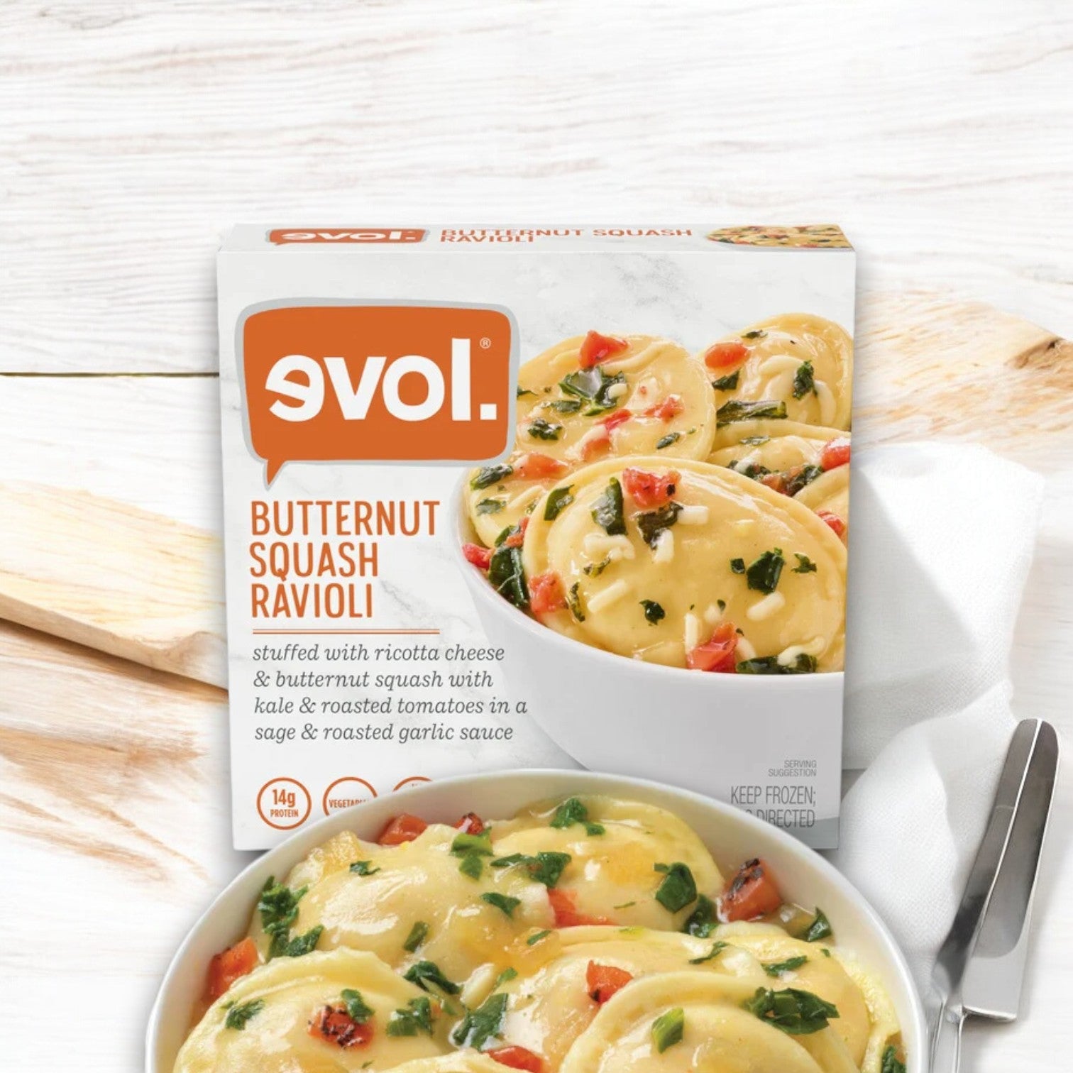 An 8.13 oz box of Evol Vegetarian Butternut Squash & Sage Ravioli, placed next to a bowl of the prepared ravioli on a white surface, garnished with kale, roasted tomatoes, and sage in a roasted garlic sauce. This single-serve meal offers a gourmet touch perfect for vegetarians.