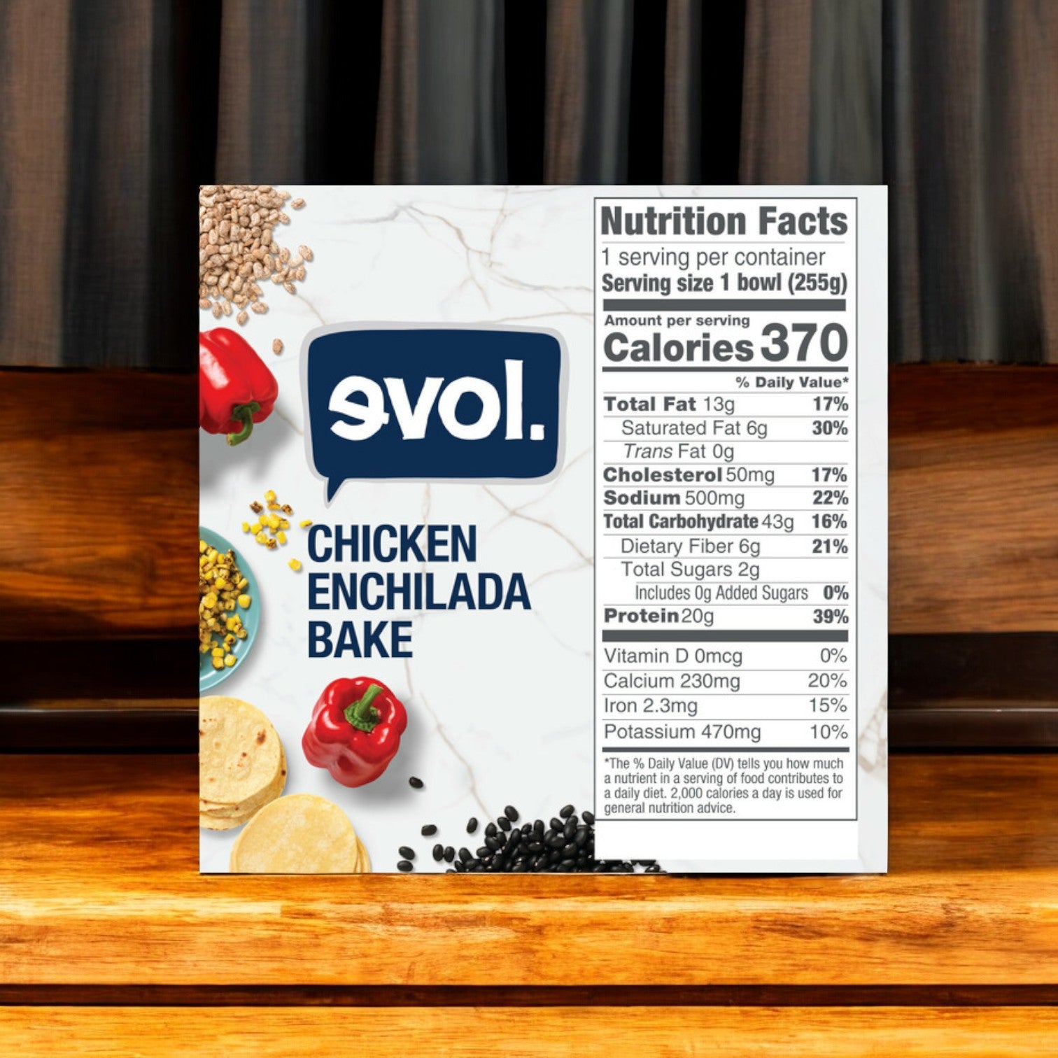 A box of Evol Chicken Enchilada Bake, Gluten Free, 9 oz. - 1 Count, is positioned on a wooden surface with a curtain backdrop. The box showcases a nutrition facts label highlighting its gourmet quality with 370 calories per serving and other nutritional details.