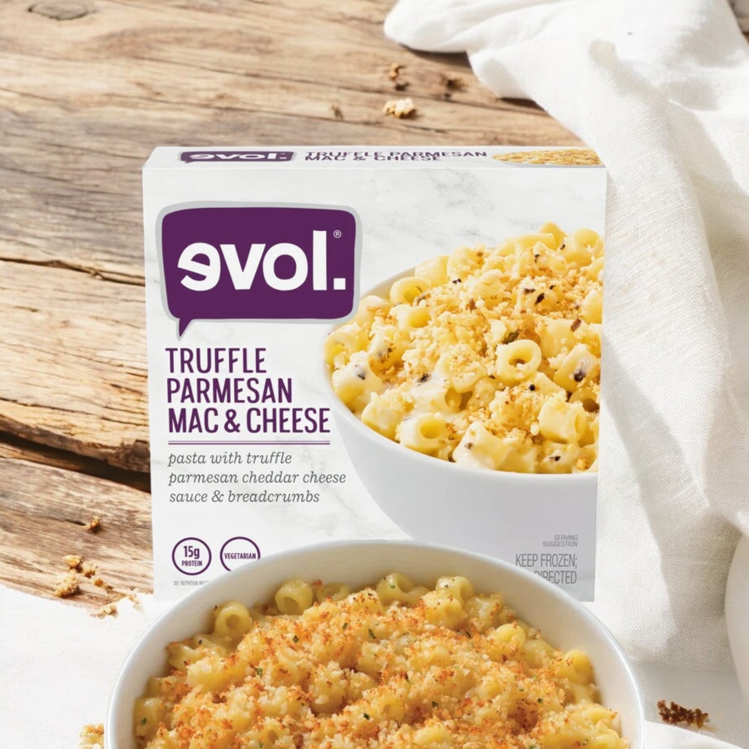 An 8 oz. box of Evol Vegetarian Truffle Parmesan Mac and Cheese - 1 Count is displayed next to a prepared bowl of the same pasta on a wooden surface, showcasing pasta in a creamy truffle-infused Parmesan cheese sauce and topped with breadcrumbs.