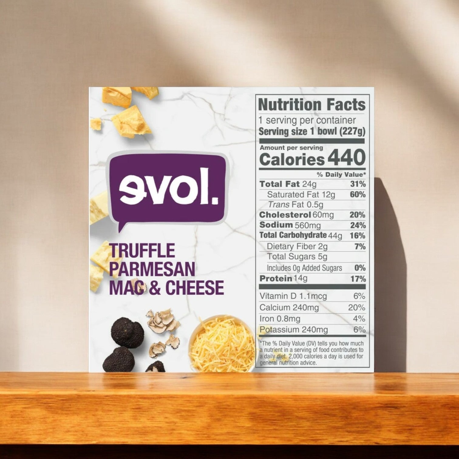 The Evol Vegetarian Truffle Parmesan Mac and Cheese, 8 oz. -1 Count box from the brand Evol is displayed on a wooden surface. Highlighted on the box is the nutrition facts panel, showing that it contains 440 calories, 24g total fat, and provides 60% of the daily recommended calcium per serving. Indulge in this gourmet mac and cheese featuring a creamy truffle-infused Parmesan cheese sauce.