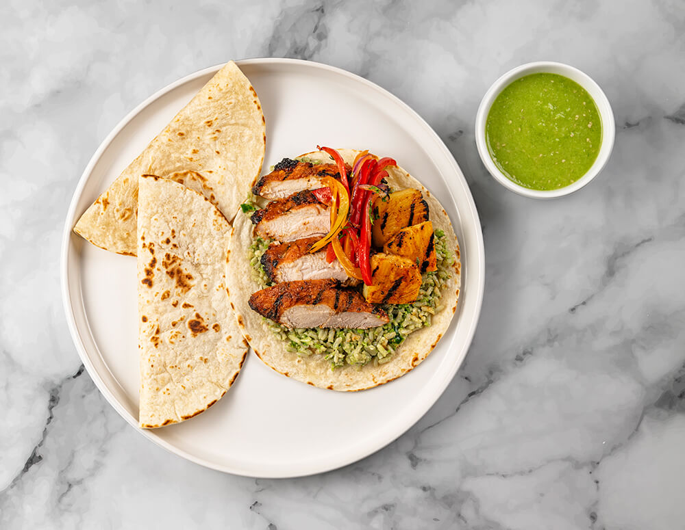 A serving of FoodHaul Al Pastor Chicken Tacos including grilled meat slices, rice, vegetables, tortillas, and a bowl of green sauce on a marble surface.