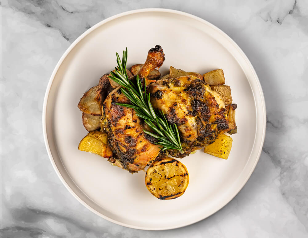 A top-down view of Food Haul's Roasted Half Brick Chicken, garnished with herbs, served alongside roasted potatoes and lemon slices.