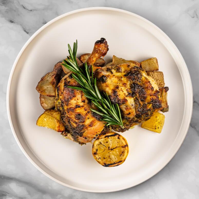 The FoodHaul Roasted Half Brick Chicken, featuring crispy skin, is accompanied by rosemary-infused potatoes and lemon slices. It is artfully presented on a round white plate against a marble background.