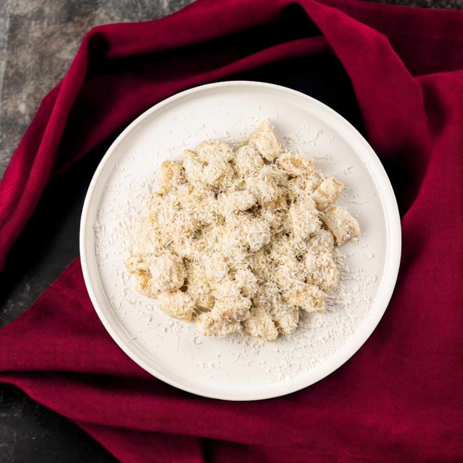 A white plate of Handmade Gnocchi Tartufo from the FoodHaul Bundle by Fabio Viviani, lavishly garnished with grated cheese, rests on a dark surface, enclosed by a vibrant red cloth.