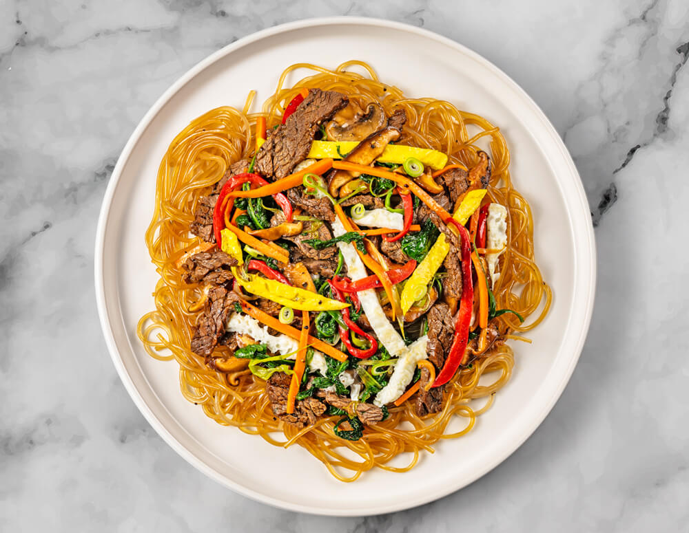 A plate of stir-fried glass noodles with slices of Food Haul's Korean Flank Steak, red and yellow bell peppers, spinach, and other vegetables.