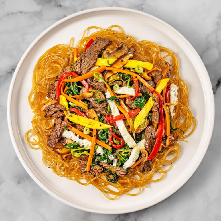 A plate of Japchae showcasing FoodHaul Korean Flank Steak, along with glass noodles, spinach, carrots, red bell peppers, and egg strips sits on a marble surface. This delightful dish is a staple in Korean cuisine and often features a savory marinade that enhances the tender beef from FoodHaul.