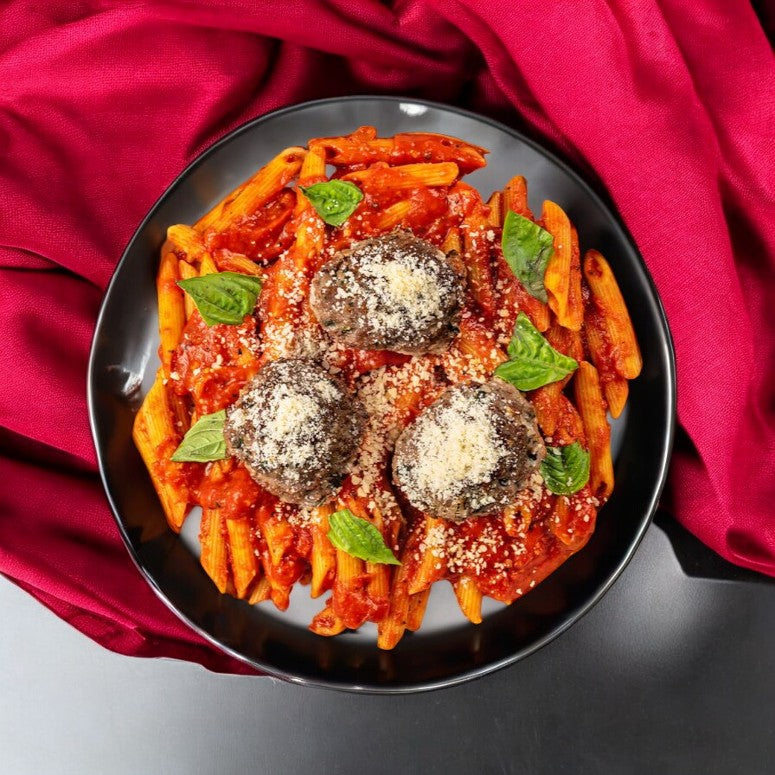 A black plate presenting a luxurious serving of penne pasta in a rich tomato sauce, topped with three generous Wagyu meatballs, grated cheese, and basil leaves. Gracefully adorned with a red cloth, this exquisite dish would be an ideal addition to any FoodHaul Bundle featuring four amazing chefs’ creations.