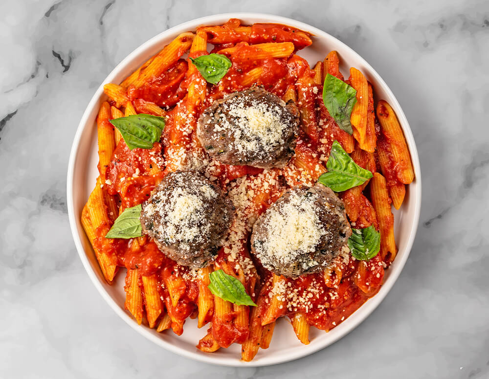 A plate of Wagyu Penne with Meatballs, a 16 oz entree by FoodHaul, features penne pasta in red sauce topped with three meatballs, grated cheese, and fresh basil leaves.