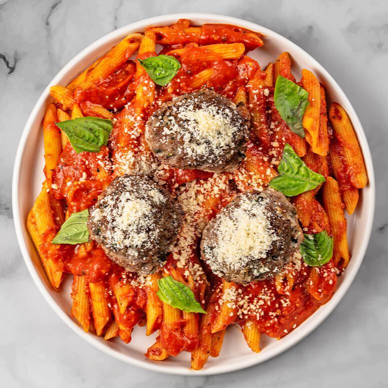 A serving of FoodHaul's Wagyu Penne With Meatballs, featuring penne pasta in rich tomato sauce, crowned with three tender Wagyu beef meatballs, grated cheese, and fresh basil leaves on a marble surface—a gourmet twist highlighting premium ingredients.