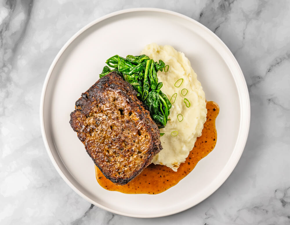 A plate featuring FoodHaul Wagyu Beef Meatloaf topped with gravy, mashed potatoes garnished with green onions, and a serving of sautéed spinach.