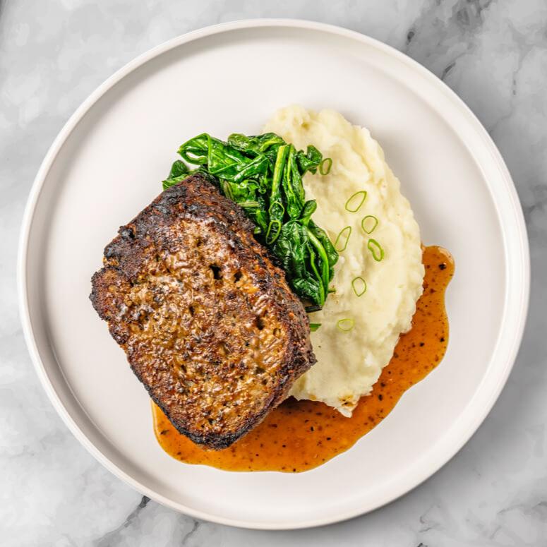 A dish featuring the FoodHaul Wagyu Beef Meatloaf, known for its tender texture, complemented by mashed potatoes garnished with green onions, sautéed spinach, and a rich brown sauce.