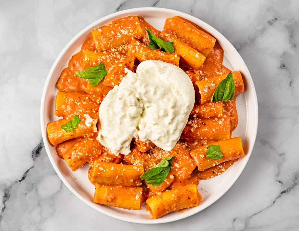 A plate of FoodHaul Spicy Rigatoni in tomato sauce from Food Haul, topped with a large portion of burrata cheese, grated cheese, and garnished with fresh basil leaves.