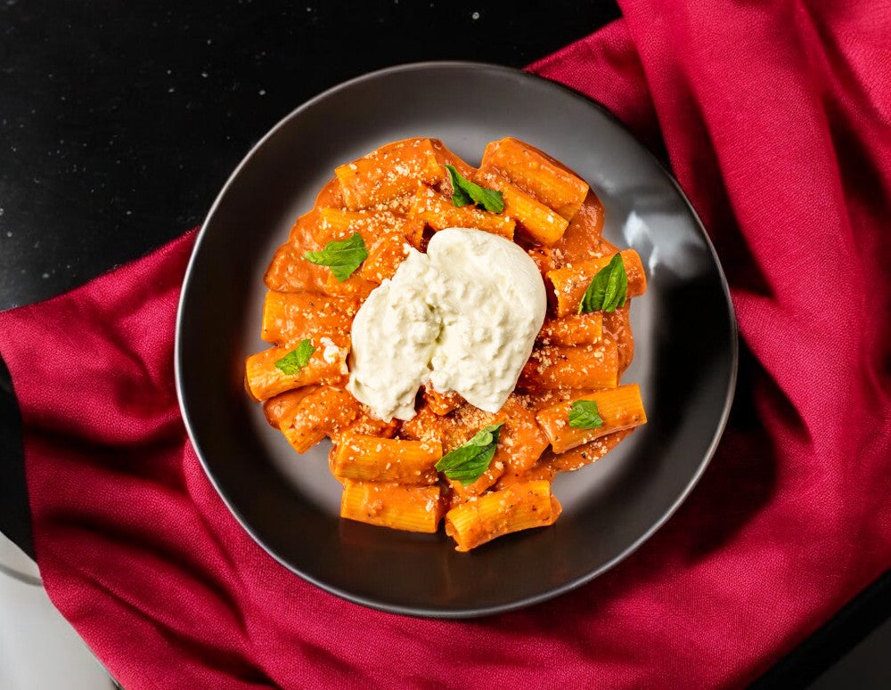 A black bowl featuring Spicy Rigatoni in a savory red sauce, adorned with a dollop of white cheese and garnished with fresh basil leaves. The bowl rests on a red cloth, perfectly enhancing your FoodHaul Bundle by Fabio Viviani for an exquisite dining experience.