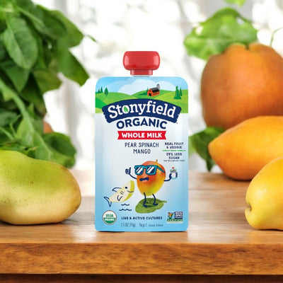 Stonyfield's Organic Pear Spinach Mango Kids' Yogurt pouch on a wooden surface with pears and mangos in the background, perfect as a kid-friendly snack.