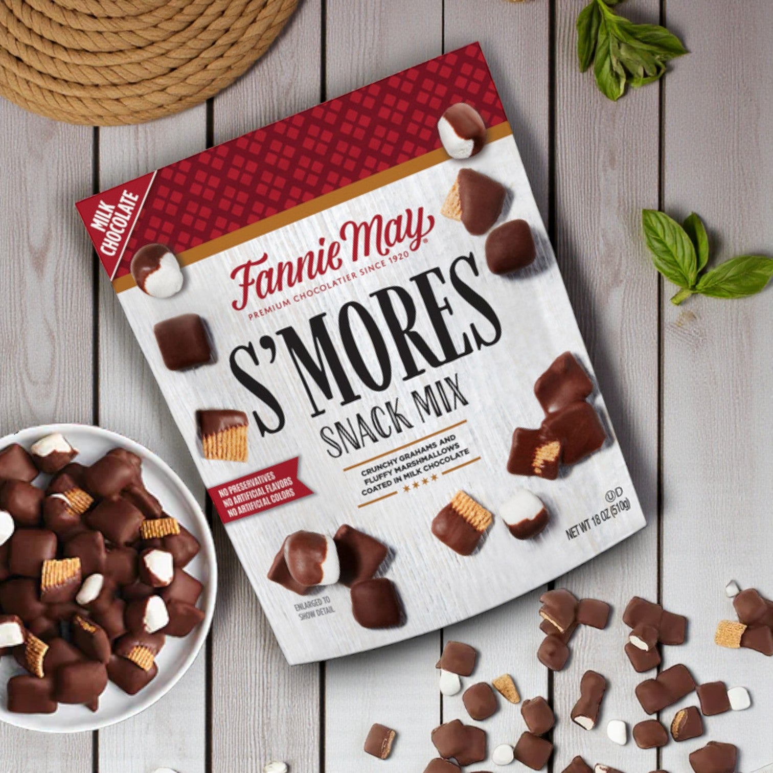 An 18 oz Fannie May S’mores Snack Mix Bag rests on a wooden surface, surrounded by gourmet chocolate treats and green plant leaves, emphasizing the classic s’mores flavor from Fannie May.