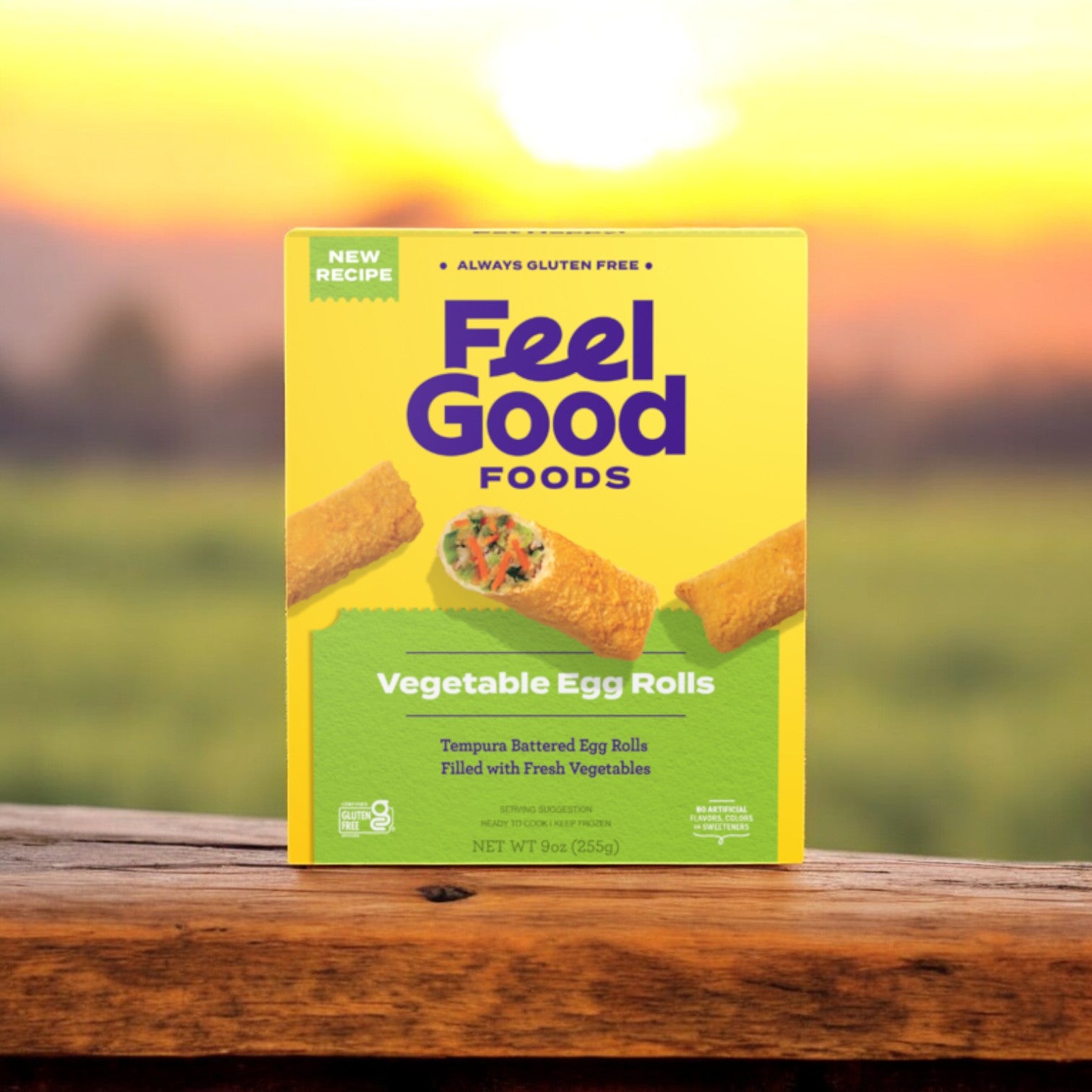 A box of Feel Good Foods Gluten-Free Vegetable Egg Rolls, 9 oz sits on a wooden surface against the backdrop of a sunset. The packaging prominently features "Always Gluten Free" and advertises a "New Recipe," making these allergen-friendly snacks ideal for anyone craving tasty, gluten-free egg rolls.