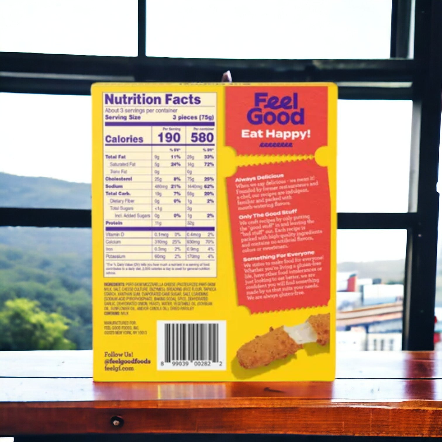 A vibrant box of Feel Good Foods Breaded Mozzarella Sticks features the "Feel Good, Eat Happy!" slogan, nutritional details, and images of the gluten-free snack on a wooden table by a window with a scenic view outside. Savor this shareable treat as part of your snacking routine!