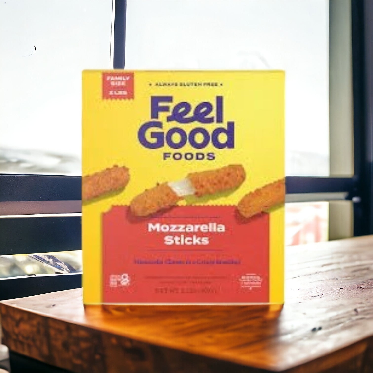 Three boxes of Feel Good Foods Gluten Free Breaded Mozzarella Sticks, the ultimate comfort food, sit invitingly on a wooden table with a window in the background. These shareable snacks promise to delight everyone at your next gathering.