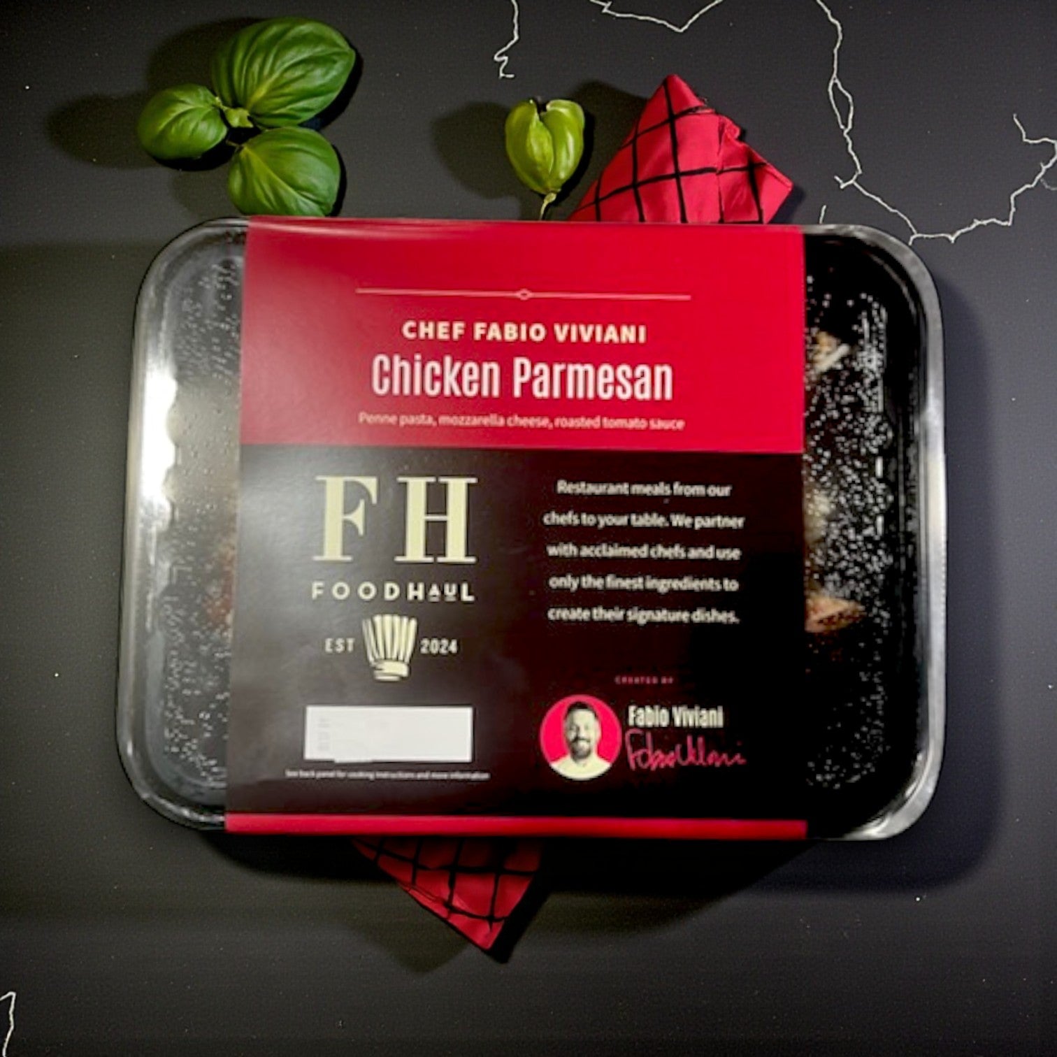 Foodhaul Frozen Meal– Chicken Parmesan by Fabio Viviani, with penne pasta, mozzarella cheese, and tomato sauce, is displayed next to fresh basil leaves and a red-patterned napkin. Brand: FoodHaul.