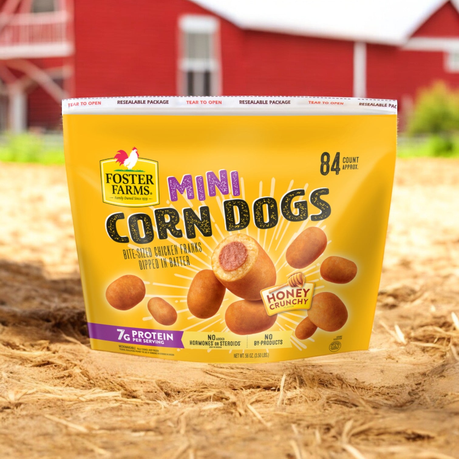 A bag of Foster Farms Corn Dogs, Mini, Honey Crunchy (84 count) is placed on a sandy surface in front of a blurred red barn, emphasizing its honey crunch flavor and protein content, along with the irresistible taste of Classic Buffalo flavor.