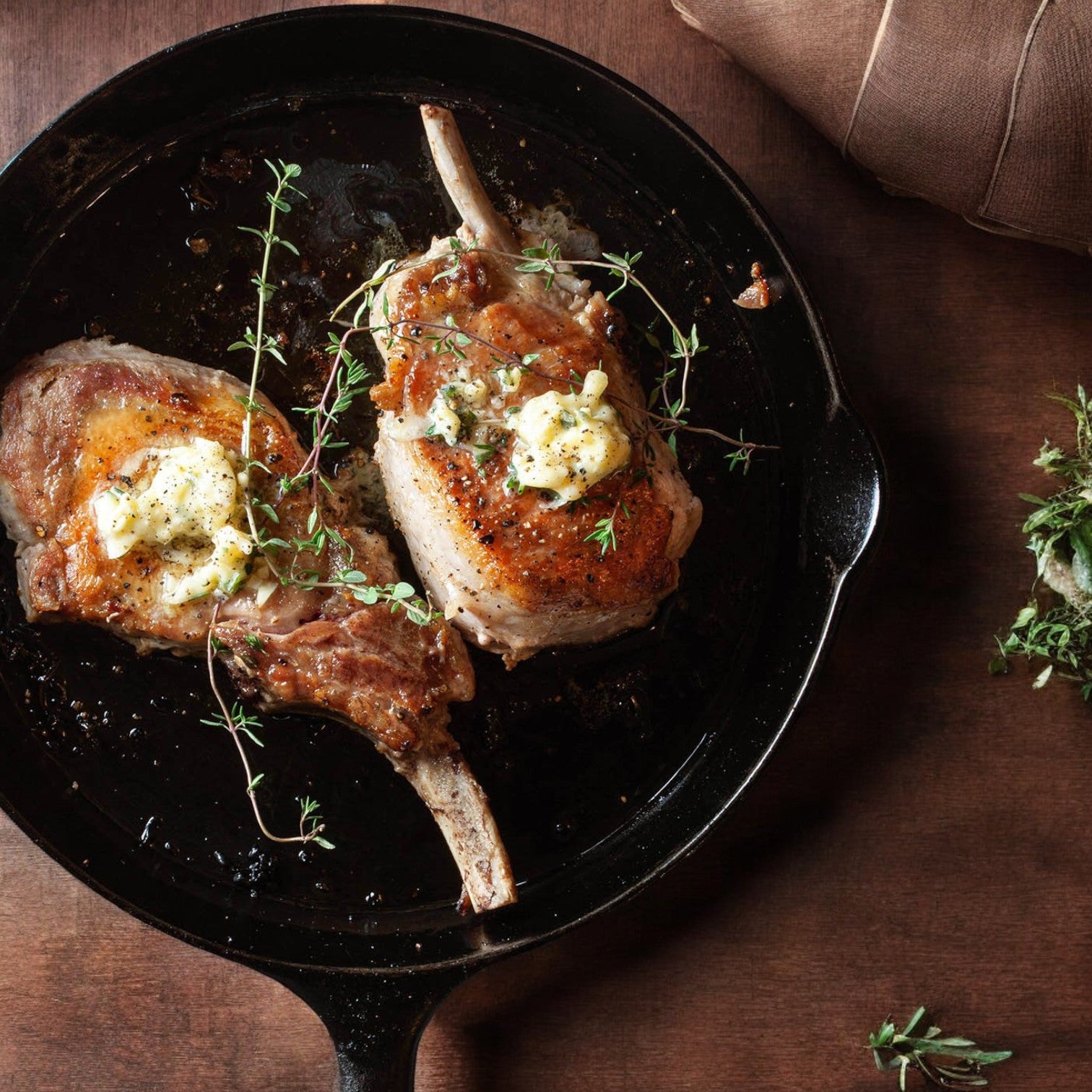 Two Mino Prime Steaks French pork chops sizzle in a skillet, adorned with fresh herbs and butter, resting on a wooden surface with a rolled napkin nearby.