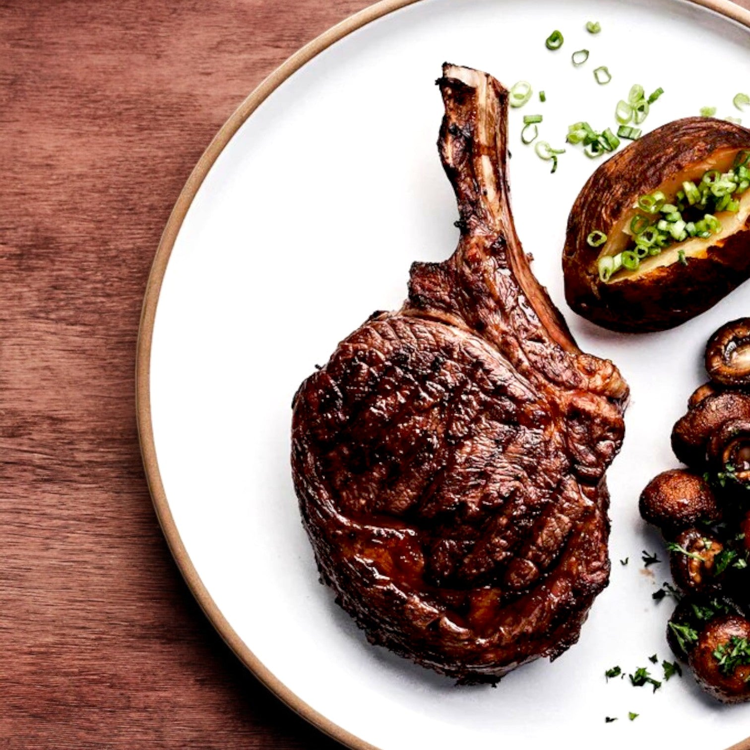 A Mino Prime Steaks, Three, 22 oz "Aleck Cut" Cowboy Ribeye is served on a round white plate with a baked potato garnished with green onions and a side of sautéed mushrooms, elegantly placed on a wooden surface.