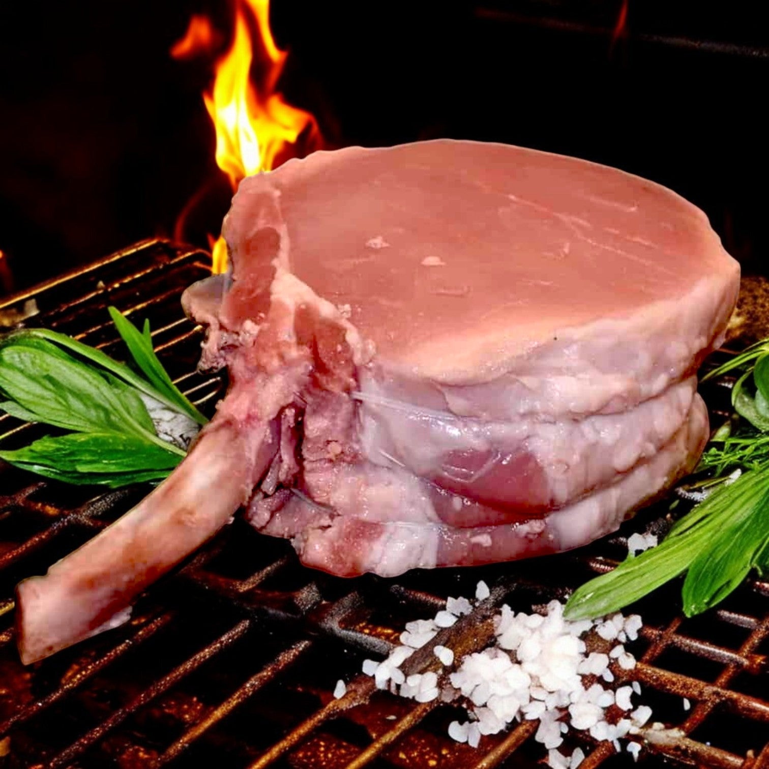 The Mino Prime Steaks 12 oz French pork chop sizzles on the grill, surrounded by herbs and coarse salt, with flames crackling in the background—a perfect high-protein feast awaiting its savory transformation.