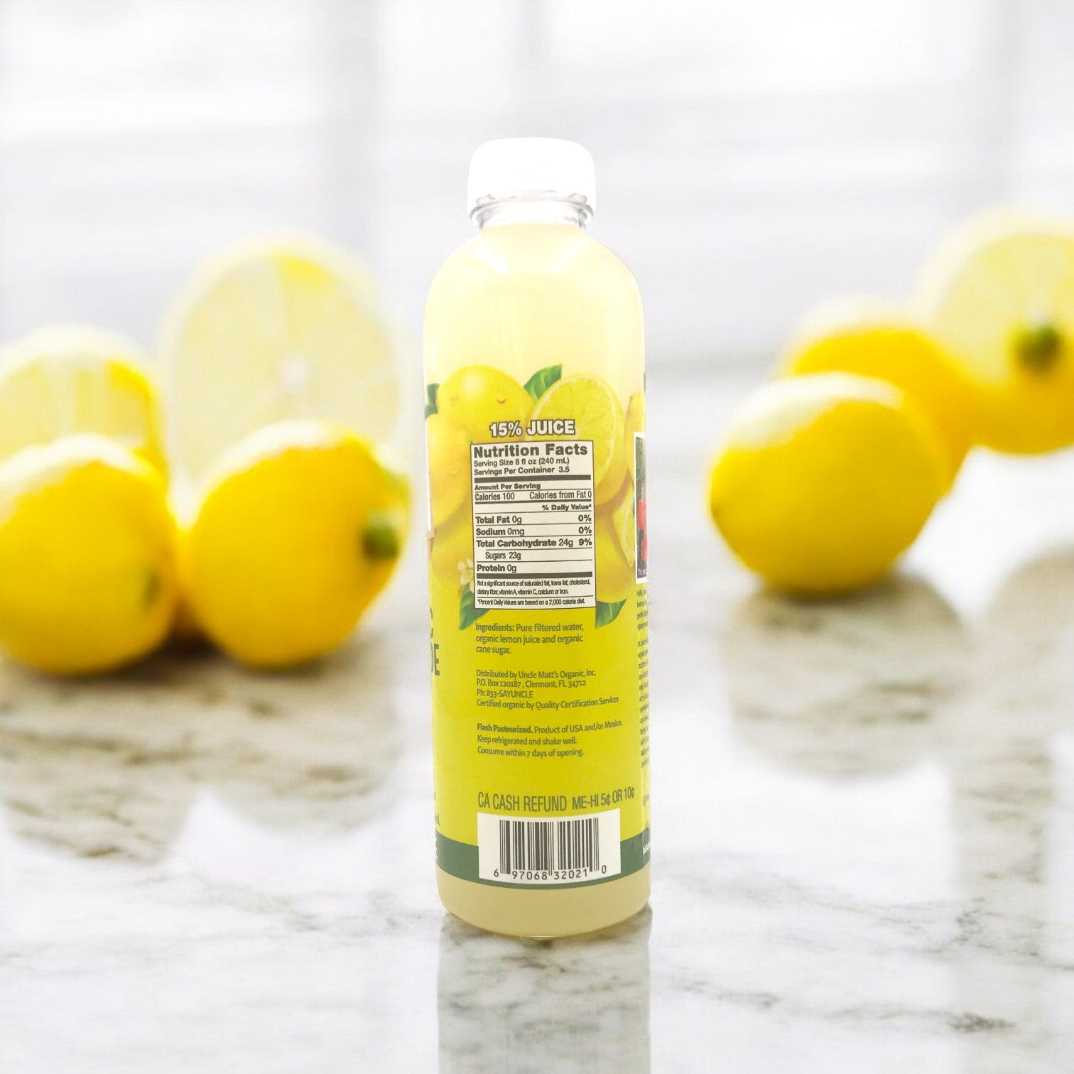 A bottle of Uncle Matt's Organic Lemonade, 12 oz, with a nutrition facts label is placed on a marble surface. Whole organic lemons are in the background, emphasizing the freshness of this No Sugar Added beverage from Uncle Matt's.