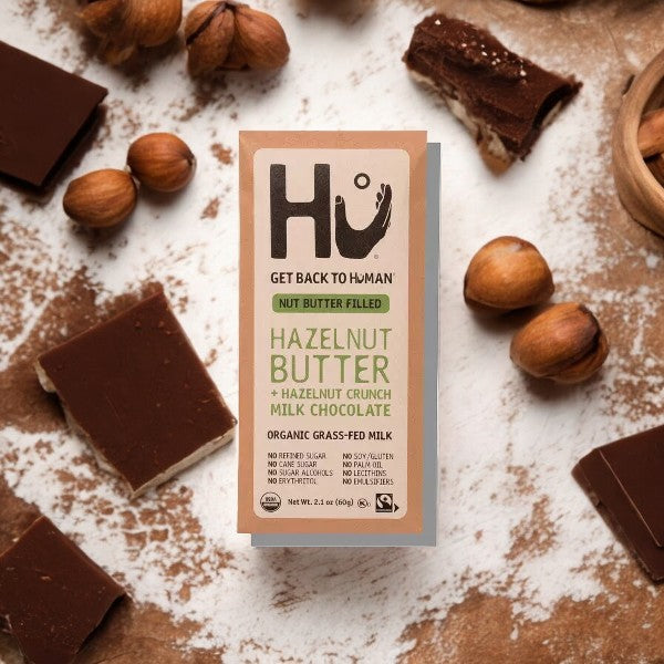 A Hu Hazelnut Butter + Hazelnut Crunch Milk Chocolate Candy bar (2.1 oz, 1 Count), made with pure ingredients, is displayed on a brown background, surrounded by luscious chocolate pieces and hazelnuts.