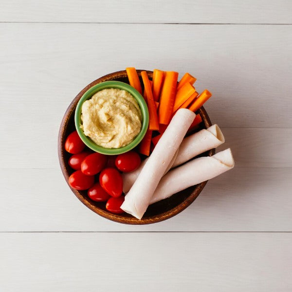 A bowl with hummus, cherry tomatoes, carrot sticks, and rolled slices of Applegate Natural Sliced Oven Roasted Turkey Breast arranged on a white wooden surface.