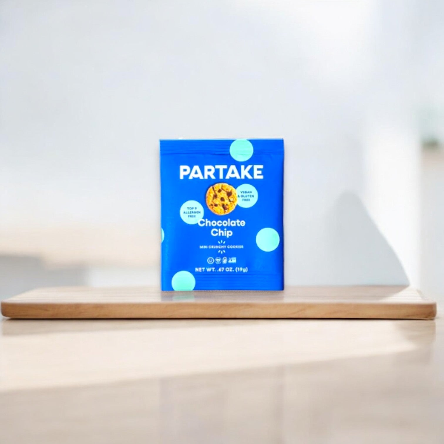 A blue package of Partake Crunchy Mini Chocolate Chip Cookie Snack Packs, 1 Count by Partake is displayed on a wooden board. The package highlights allergen-friendly, vegan, and gluten-free labels, making it the perfect addition to your snack packs.