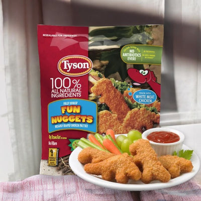 A package of Tyson Fully Cooked Fun Chicken Nuggets is displayed in the background with a plate of whole grain breaded, dinosaur-shaped nuggets, a small bowl of ketchup, and grapes in the foreground.