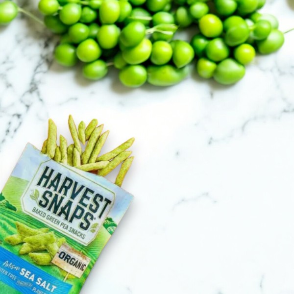 Packet of Harvest Snaps Organic Artisan Sea Salt Baked Green Pea Snacks - 3 oz on a marble surface with fresh green peas in the background, these delicious veggie crisps are packed with plant protein.