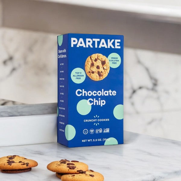 A box of Partake Gluten Free Vegan Chocolate Chip Cookies is displayed on a marble countertop with a few cookies in front of it. The packaging highlights that these cookies are vegan, gluten-free, allergen-free, and emphasizes sustainability and community support in every bite.