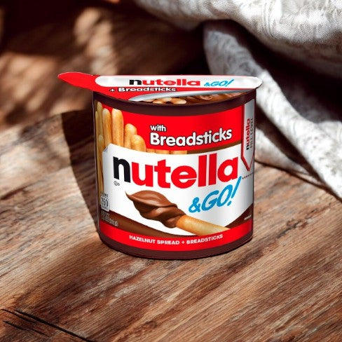 A container of Nutella and Go Breadsticks - 1.8 oz - 1 count rests on a wooden surface, partially illuminated by sunlight. The red and white packaging from Nutella prominently showcases the mouthwatering hazelnut spread paired with crispy breadsticks, inviting you to dip into indulgence.