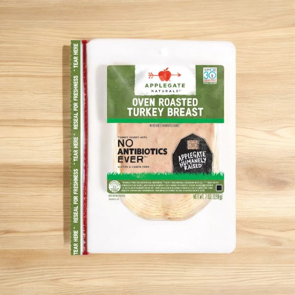 A package of Applegate Natural Sliced Oven Roasted Turkey Breast, 7 oz- 1 Count, sits on a wooden surface. The product, from the brand Applegate, is labeled "No Antibiotics Ever," "Humanely Raised," and "Whole30 Approved," with a reseal strip visible on the left side.