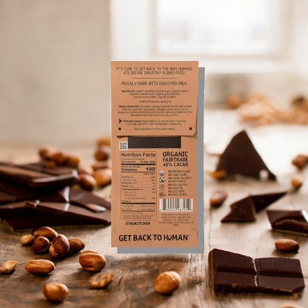 A Hu Hazelnut Butter + Hazelnut Crunch Milk Chocolate Candy bar stands on a table surrounded by scattered hazelnuts and broken chocolate pieces. The back of the 2.1 oz bar displays nutrition facts and pure ingredients, with a message to "Get Back to Human." This organic Hu milk chocolate treat also features a delightful hazelnut butter center.