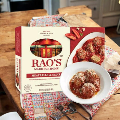 A box of Rao's Made For Home Family Size Frozen Meatballs and Sauce by Easy Lunches is displayed beside a white bowl containing three meatballs garnished with grated cheese, all set on a patterned cloth atop a wooden table.