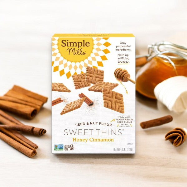 A box of Simple Mills Honey Cinnamon Sweet Thins made from Seed & Nut Flour is displayed with cinnamon sticks, a small bowl of honey, and a honey dipper in the background.