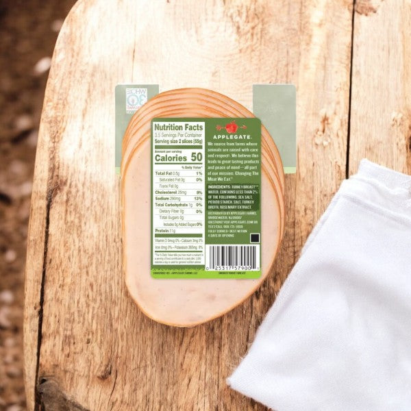 A packaged slice of Applegate Natural Smoked Turkey Breast, 7 oz- 1 Count, on a wooden table. The green label displays nutritional information, including 50 calories per serving, and brand details. A white napkin is partially visible, highlighting the antibiotic-free and fully cooked nature of the product.