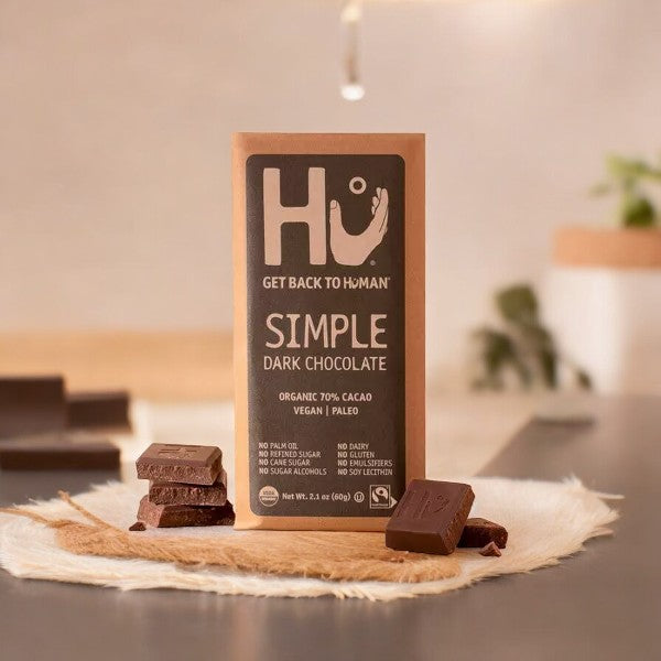 A package of "Hu Simple Dark Chocolate 70% Cacao Candy" is on a tabletop. Several pieces of the chocolate bar are scattered in front of the package, which is labeled organic, vegan, paleo, and made with organic unrefined coconut sugar.