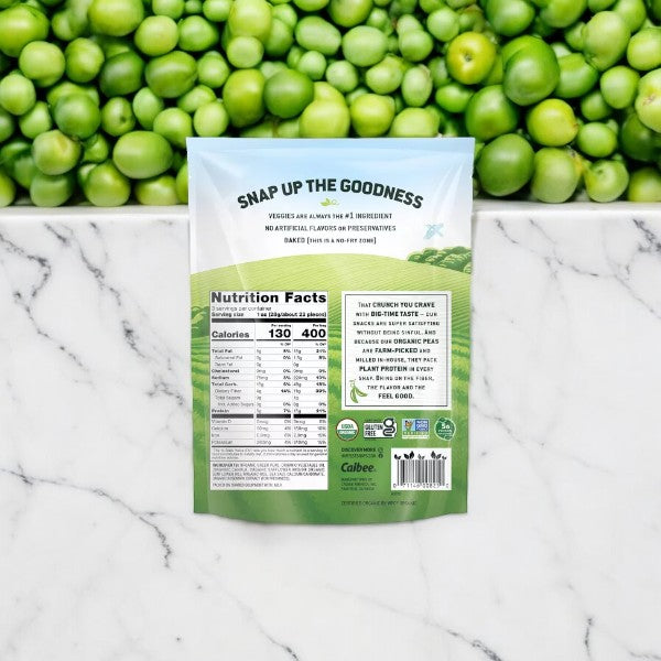 A 3 oz package of Harvest Snaps Organic Artisan Sea Salt Baked Green Pea Snacks is set on a marble surface, with the nutritional information displayed on the back and fresh peas in the background.