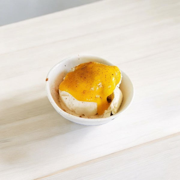 A bowl of vanilla ice cream topped with a vibrant sauce made from Golden Farms Organic Mango Squeeze and a sprinkle of spices, all on a light wooden surface.