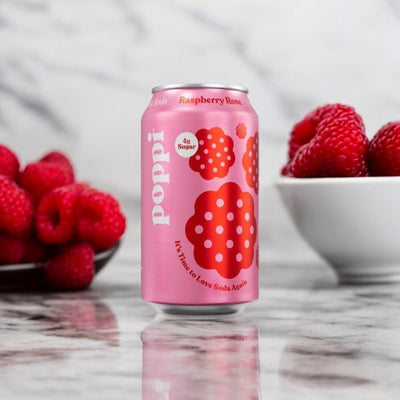 A Poppi Prebiotic Soda, Raspberry Rose, 1-12 fl oz Can sits on a marble countertop next to bowls filled with fresh raspberries, showcasing this low-calorie beverage packed with prebiotics.