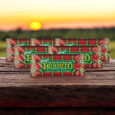 Several packages of Amy's Burrito 6 Pack—featuring 6 Burritos with Cheddar Cheese—are displayed on a wooden surface with a sunset in the background.