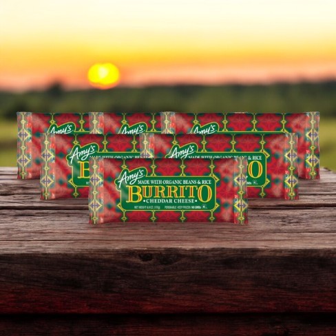 Several packages of Amy's Burrito 6 Pack—featuring 6 Burritos with Cheddar Cheese—are displayed on a wooden surface with a sunset in the background.