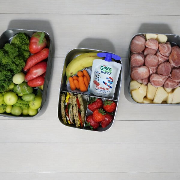 Three metal containers are filled with various food items. The left container has fruits and vegetables, the middle offers a nutritious snack with a banana, carrots, strawberries, and a GoGo squeeZ® yogurtZ - Strawberry 3 oz pouch, while the right contains sliced meat and cheese.