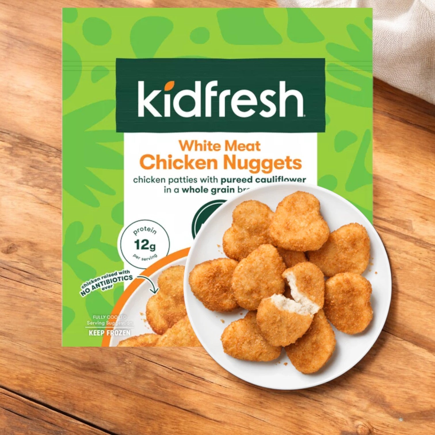 A packaging of Kidfresh Frozen Chicken Nuggets - 16.35 oz is shown next to a plate with heart-shaped, white-meat nuggets, all on a wooden surface. These nuggets are also packed with hidden veggies for added nutrition.