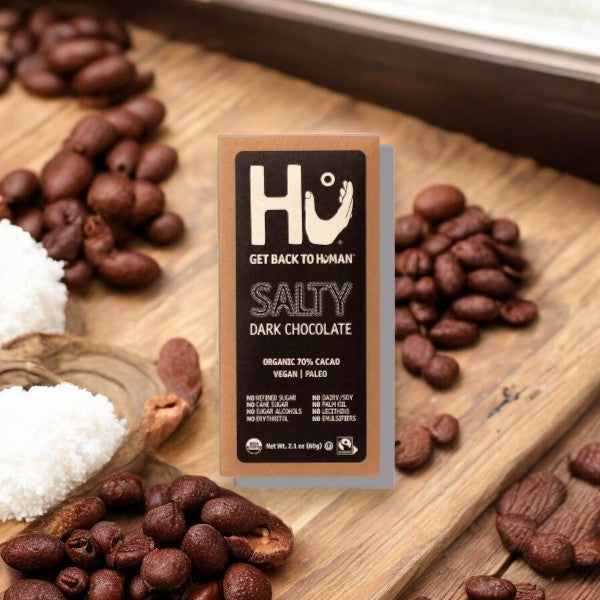 A bar of Hu Salty Dark Chocolate 70% Cacao Candy (2.1 oz, 1 Count) from Hu is placed on a wooden surface with coffee beans, salt, and coconut flakes surrounding it. Made with organic ingredients, this salty chocolate bar promises a rich and flavorful experience.