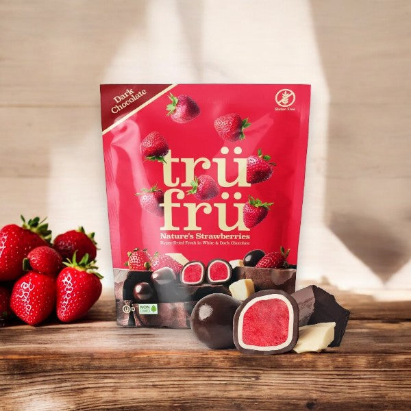 The red packaging of Tru Fru Chocolate Covered Strawberry's- 4.2 oz - 1 Count is placed on a wooden surface. Several fresh strawberries and pieces of the dark chocolate-coated strawberries are also displayed, making it perfect for convenient snacking.